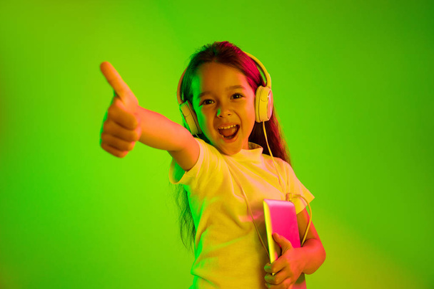 Portrait of little girl in headphones on green background in neon light - Foto, afbeelding