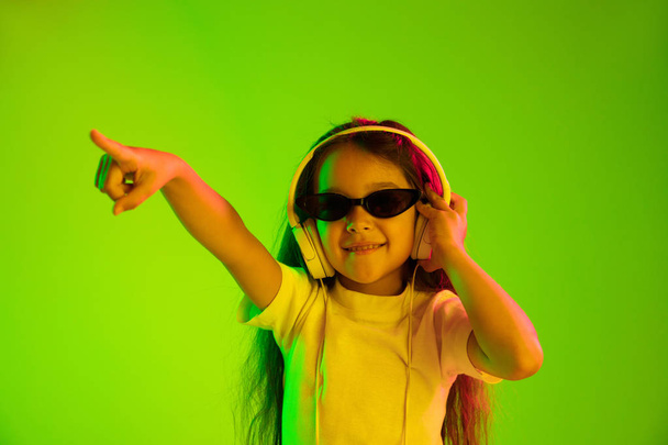 Portrait of little girl in headphones on green background in neon light - Φωτογραφία, εικόνα