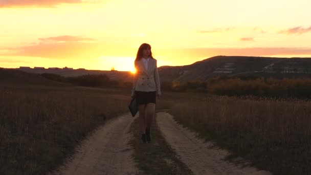 太陽の手でブリーフケースの国の道路に沿って歩くセクシーなビジネスウーマンは日没でゆらめきます。田舎で働くセクシーなビジネス女性。美しい少女は、田舎を旅します。. - 映像、動画