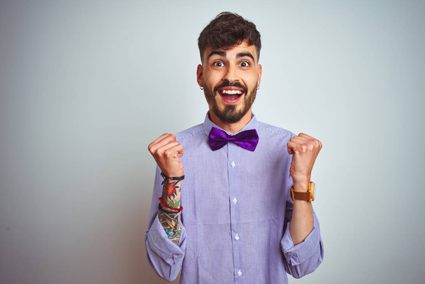 紫色のシャツと蝶ネクタイを着たタトゥーを持つ若い男は、腕を上げて目を開けて成功に驚き、驚きました。勝者のコンセプト. - 写真・画像