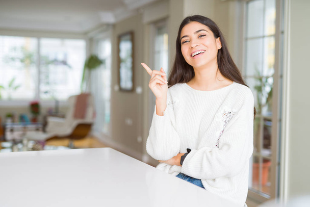Young beautiful woman at home on white table with a big smile on face, pointing with hand and finger to the side looking at the camera. - Photo, Image