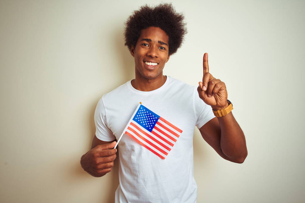 Афро-человек с флагом Соединенных Штатов Америки, стоящий на изолированном белом фоне, удивленный идеей или вопросом, указывая пальцем со счастливым лицом, номер один
 - Фото, изображение