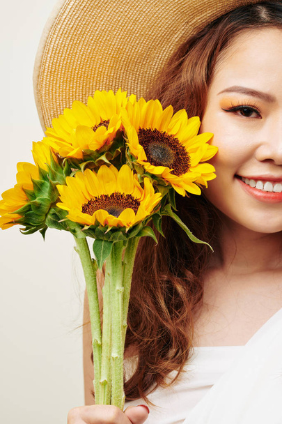 Portrét krásné asijské ženy v klobouku držící slunečnice a usmívající se na kameře  - Fotografie, Obrázek