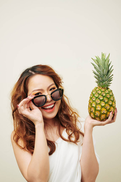 Portrait of Asian beautiful woman with red curly hair wearing sunglasses holding pineapple and smiling at camera over white background - Photo, Image