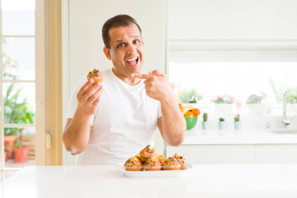 Middle age man eating chocolate chip muffins at home very happy pointing with hand and finger - Photo, Image