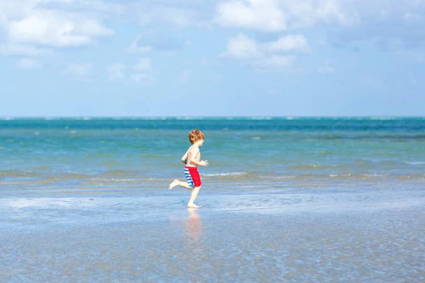Niño activo divirtiéndose en la playa de Miami, Key Biscayne. Feliz niño lindo corriendo cerca del océano en un día cálido y soleado. Niño sano en pantalones de baño cazando aves gaviotas - Foto, Imagen