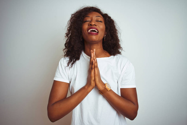 Młoda African American kobieta nosi t-shirt stojący na białym tle białe żebractwa i modlitwy z rąk wraz z nadzieją wyraz na twarzy bardzo emocjonalny i zaniepokojony. Prosząc o przebaczenie. Koncepcja religii. - Zdjęcie, obraz