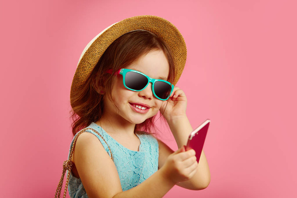Little girl in summer clothes, wearing a beach hat, sunglasses, looking at the phone, stands on pink isolated background. - Foto, afbeelding