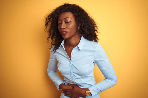 African American zakenvrouw dragen van elegante shirt over geïsoleerde gele achtergrond met hand op maag omdat indigestie, pijnlijke ziekte gevoel onwel. Ache concept. - Foto, afbeelding