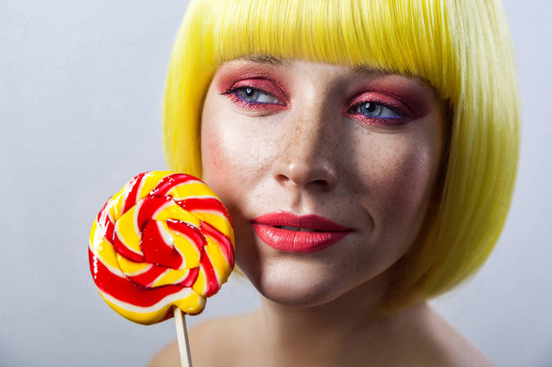 Beauty portrait of cute young woman with freckles and red makeup in yellow wig holding colorful candy stick near face and looking aside with passion  - Photo, image