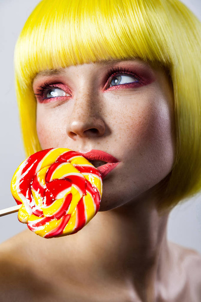 Beauty portrait of cute young woman with freckles and red makeup in yellow wig holding colorful candy stick near lips and looking aside with passion  - Foto, afbeelding