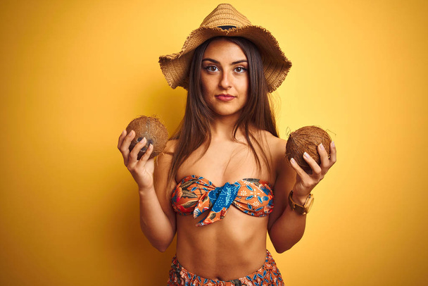 Woman on vacation wearing bikini and hat holding coconut over isolated yellow background with a confident expression on smart face thinking serious - Foto, Imagen