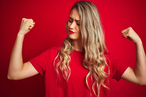 Mujer hermosa joven con camiseta básica de pie sobre fondo rojo aislado que muestra los músculos de los brazos sonriendo orgulloso. Concepto Fitness
. - Foto, imagen