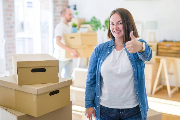 Young couple arround cardboard boxes moving to a new house, plus size woman standing at home doing happy thumbs up gesture with hand. Approving expression looking at the camera with showing success. - Фото, зображення