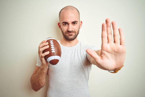 Young man holding rugby american football ball over isolated background with open hand doing stop sign with serious and confident expression, defense gesture - Photo, Image
