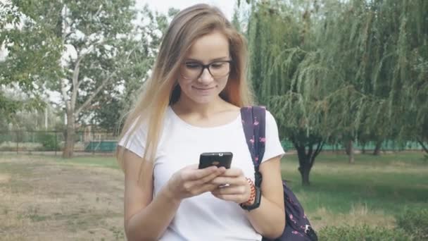 Portrait of relaxed young lady in glasses in a summer park reading a text message on her mobile phone. Beautiful young girl with blond long hair using her cell phone, outdoor. - 映像、動画