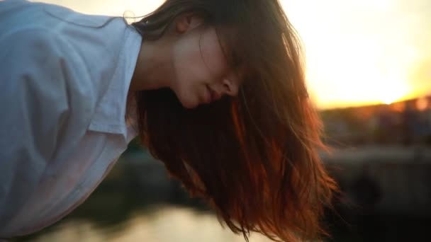 Een jonge trieste vrouw staat, over en kijkt naar het water met losse haren in wit overhemd bij zonsondergang. - Video