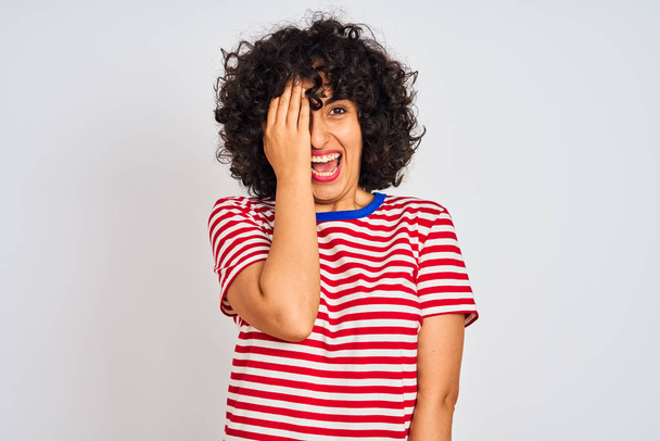 Jeune femme arabe aux cheveux bouclés portant un t-shirt rayé sur fond blanc isolé couvrant un œil avec la main, sourire confiant sur le visage et émotion surprise
. - Photo, image