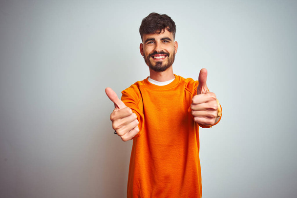 Молодой человек с татуировкой в оранжевом свитере стоит на изолированном белом фоне, одобряя позитивный жест рукой, улыбается и радуется успеху. Жест победителя
. - Фото, изображение