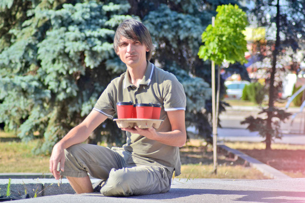 Молодой белый человек сидит на бетонном парапете в парке и держит четыре красных бумажных стакана с кофе на фоне города
 - Фото, изображение