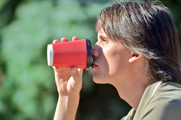 Portraite nuori valkoinen mies tumma tukka vihreä paita juo kahvia punainen paperi cup puistossa avoimin silmin
 - Valokuva, kuva