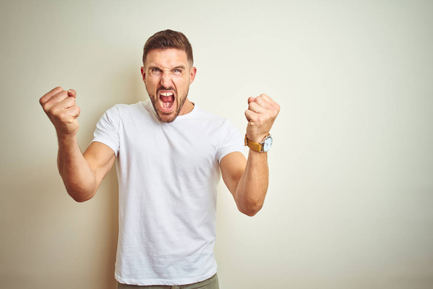 Young handsome man wearing casual white t-shirt over isolated background angry and mad raising fists frustrated and furious while shouting with anger. Rage and aggressive concept. - Foto, Bild