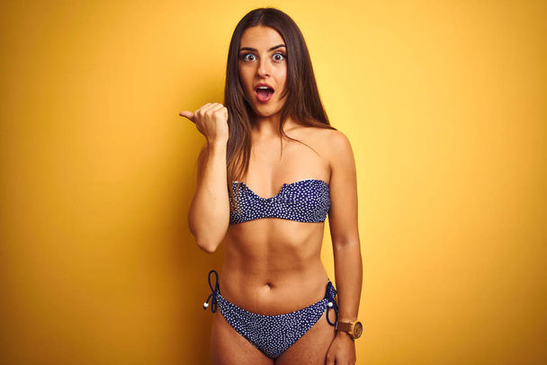 Young beautiful woman on vacation wearing bikini standing over isolated yellow background Surprised pointing with hand finger to the side, open mouth amazed expression. - Fotó, kép