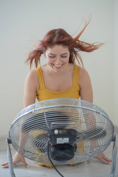 Portrait of a beautiful red-haired woman in a mustard jumpsuit enjoying the cooling breeze from a large electric fan. A smiling girl freshens up in the hot summer heat. Hair develops from the wind - Fotó, kép