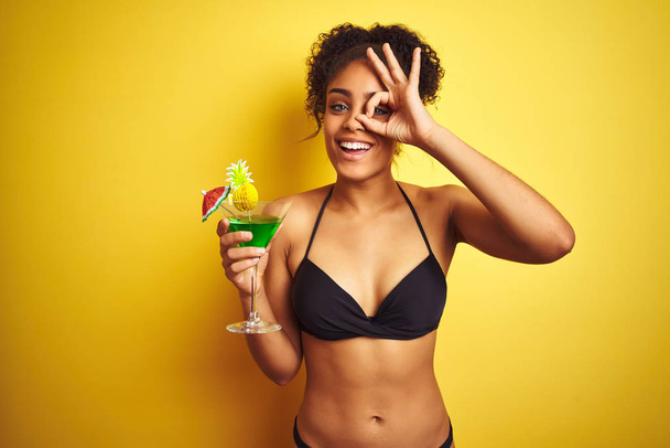 Afro vrouw op vakantie dragen bikini drinken van een cocktail over geïsoleerde gele achtergrond met gelukkig gezicht glimlachend doen OK teken met de hand op oog kijken door vingers - Foto, afbeelding