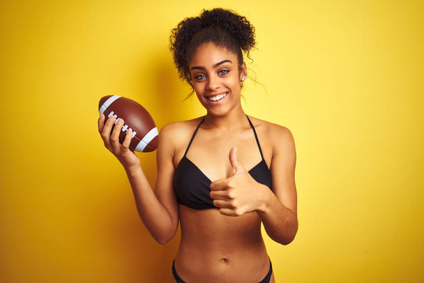 Afro american woman on vacation wearing bikini playing rugby over isolated yellow background happy with big smile doing ok sign, thumb up with fingers, excellent sign - Photo, Image