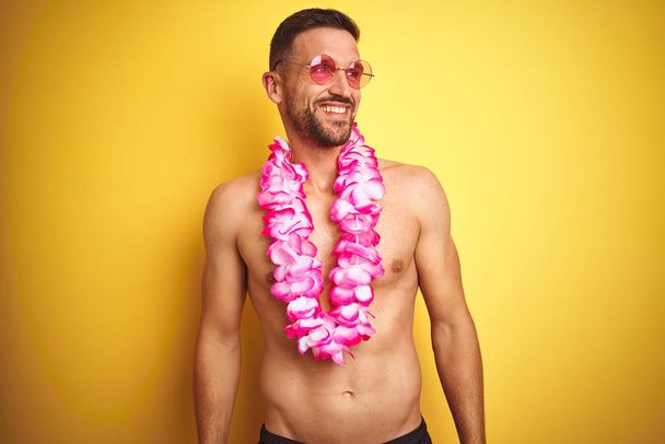 Jeune homme beau torse nu portant des lunettes de soleil et lei hawaïen rose sur fond jaune regardant de côté avec le sourire sur le visage, expression naturelle. Rire confiant
. - Photo, image