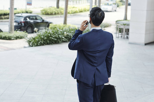 Rear view of young businessman standing outdoors and talking on phone when waiting for taxi - Photo, image