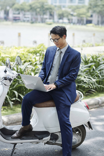 Красивый молодой азиатский предприниматель в очках и классическом костюме сидит на открытом воздухе и работает на ноутбуке
 - Фото, изображение