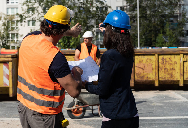 Back turned woman civil engineer discussing about plans, blueprints and paperworks with the foreman on construction site. In the background, another building contractor. Selective focus - Photo, Image