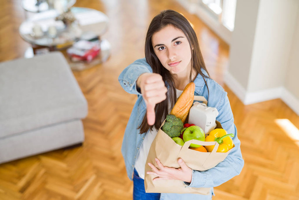 Beautiful young woman holding paper bag full of healthy groceries with angry face, negative sign showing dislike with thumbs down, rejection concept - Valokuva, kuva