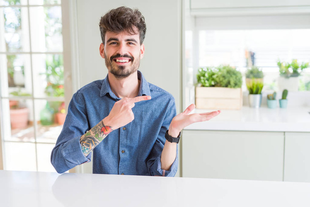 Fiatal férfi visel alkalmi póló ül a fehér asztalon meghökkent, és mosolyogva a kamerát, miközben bemutatja a kezét, és mutatva ujjal. - Fotó, kép