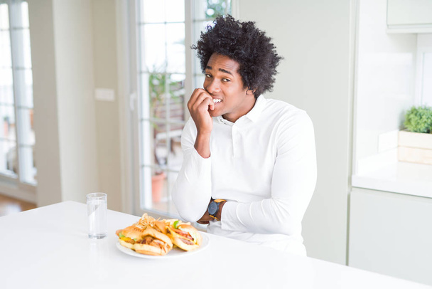 Голодный афроамериканец ест гамбургер на обед, выглядит напряженным и нервным с руками на губах, грызущими ногти. Проблема тревоги
. - Фото, изображение