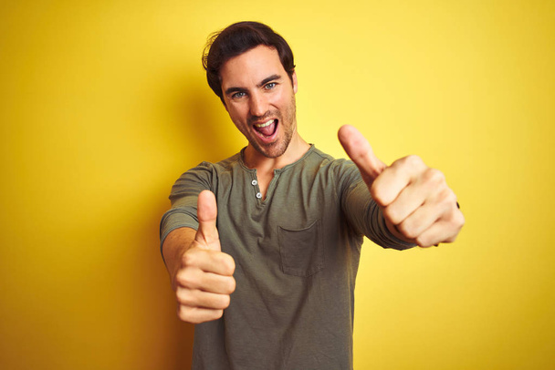 Young handsome man wearing casual t-shirt standing over isolated yellow background approving doing positive gesture with hand, thumbs up smiling and happy for success. Winner gesture. - Photo, Image