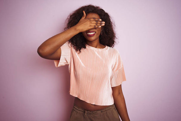Jonge Afro-Amerikaanse vrouw dragen t-shirt staande over geïsoleerde roze achtergrond glimlachen en lachen met de hand op gezicht bedekt ogen voor verrassing. Blind concept. - Foto, afbeelding