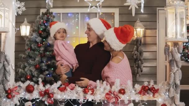Young happy family of mother, father and little daughter dressed in Santa Claus hat hugging at background of Christmas and New Year tree at porch. Family, Xmas concept in slow motion. Medium shot - Filmati, video