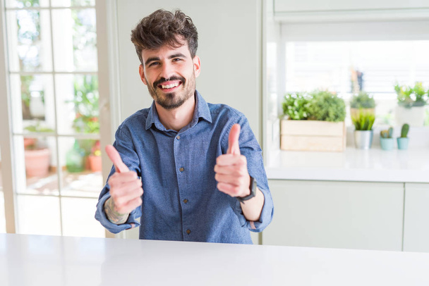 Młody mężczyzna ubrany casual shirt siedzi na białym stole zatwierdzania robi pozytywny gest z ręki, kciuki się uśmiechnięty i szczęśliwy dla sukcesu. Patrząc na aparat, gest zwycięzcy. - Zdjęcie, obraz