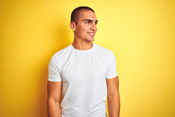 Homem caucasiano jovem vestindo t-shirt branca casual sobre fundo isolado amarelo olhando para o lado com sorriso no rosto, expressão natural. Rindo confiante
. - Foto, Imagem