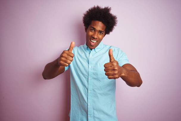 Fiatal amerikai férfi afro hajú viselt kék inget állva rózsaszín háttér jóváhagyásáról csinál pozitív gesztus kézzel, remek mosolygó és boldog a sikerhez. Győztes kézmozdulat. - Fotó, kép