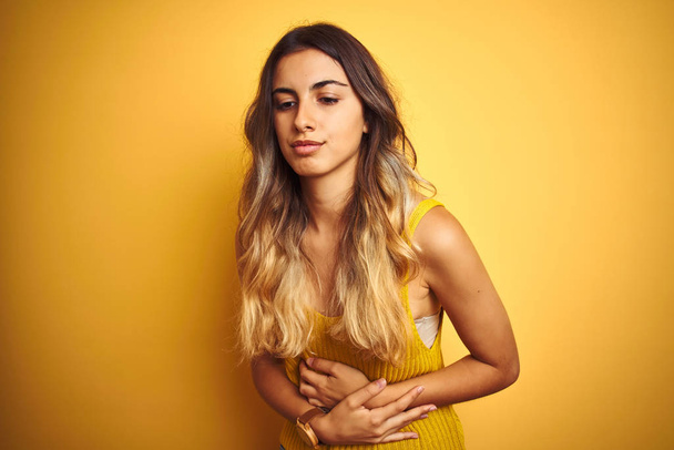 junge schöne Frau trägt T-Shirt über gelbem isolierten Hintergrund mit der Hand auf dem Bauch, weil Verdauungsstörungen, schmerzhafte Krankheit fühlen sich unwohl. Schmerzkonzept. - Foto, Bild