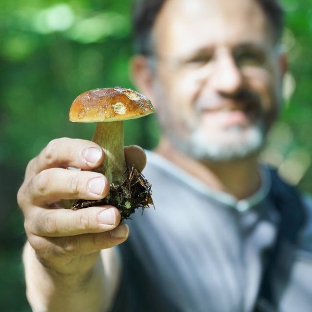 Bearded man shows a mushroom boletus edulis in hand in the forest - Photo, Image