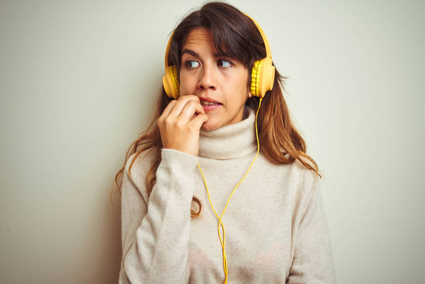 Young beautiful woman listening to music using headphones over white isolated background looking stressed and nervous with hands on mouth biting nails. Anxiety problem. - Photo, image