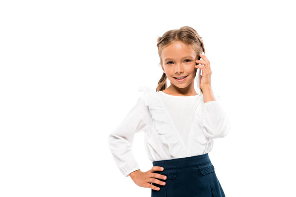 positive kid standing with hand on hip and talking on smartphone isolated on white  - Photo, Image