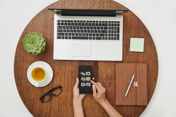 Nahaufnahme einer Frau, die mit Laptop und Notizblock an einem Holztisch sitzt und mit ihrem Handy online Imbiss bestellt - Foto, Bild