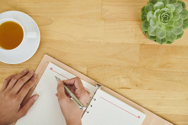 Gros plan des mains féminines tenant un stylo et des plans d'écriture dans un bloc-notes assis à une table en bois et buvant du thé
 - Photo, image