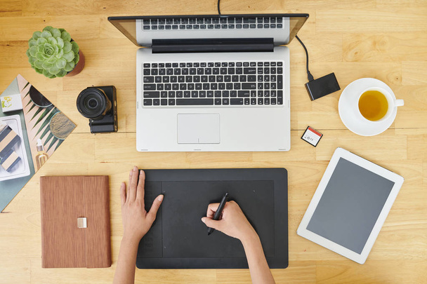 Крупный план дизайнера, сидящего за деревянным столом перед ноутбуком и использующего графический планшет в своей работе в офисе
 - Фото, изображение
